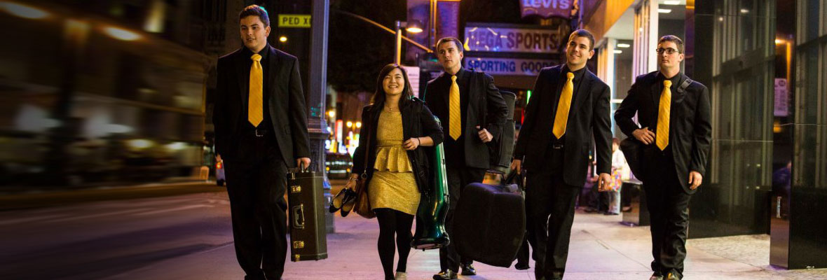 Brass players dressed up in black and gold walking at night on a city sidewalk as cars speed by.