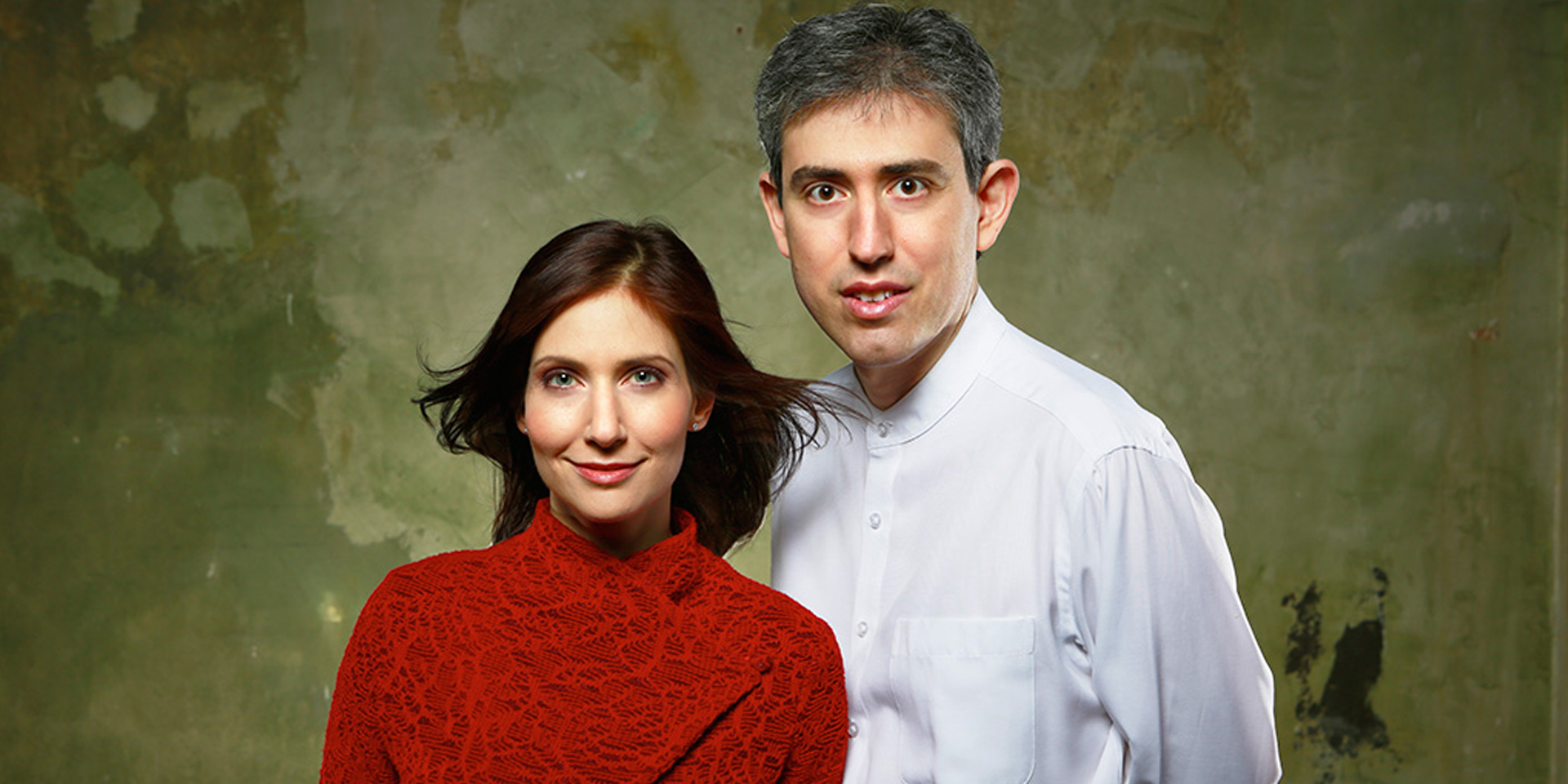 Portrait of Sivan Silver and Gil Garburg in red and white garments before a distressed wall.